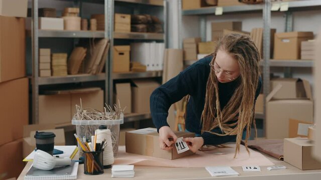 Cautiously, fragile. Attentive caucasian woman with dreadlocks wearing glasses putting special sticker while working at the warehouse. Post service and small business concept.