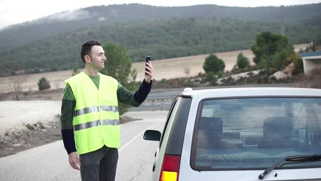 Driver in videoconference call with insurance help center. Yellow reflective vest. Emergency light V16 beacon on damaged vehicle roof. Help flash, mandatory to replace triangles in Europe. DGT Spain