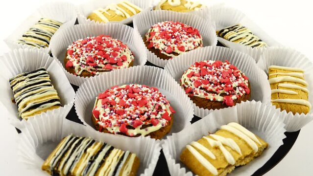 Shortbread cookies decorated with white and black chocolate icing, and red sugar sprinkles in the form of hearts in a paper molds for packaging are on a black photography turntable