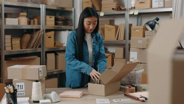 Waist up portrait view of the asian woman standing at the warehouse and putting filling at the cupboard box while packing parcels at the wooden table. Small business concept.