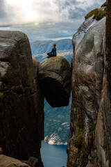 Visiting Norway Kjeragbolten located south of Lysefjorden Young woman sits on top of gian boulder...