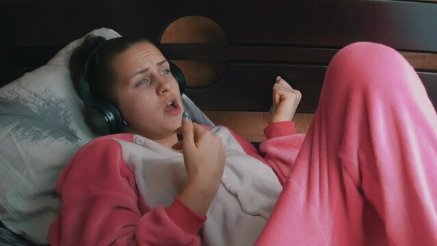 Girl in pajamas on the bed listens to music through headphones and dances. Sitting cross-legged on a hotel bed. Happy woman spends time in the evening in bed. Modern technologies and youth culture.
