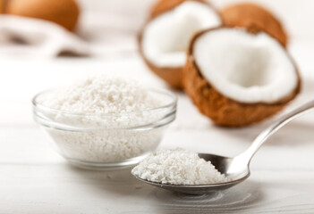 Coconut flakes in a glass bowl and spooned on a background with an open white-fleshed coconut. Composition on white texture wood. Organic healthy diet product.Side view.