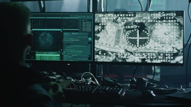 A hacker in glasses at a computer with software, during a cyber attack on the docking of the space module to the international space station in dark refuge. Footage on the screeen furnished by NASA.