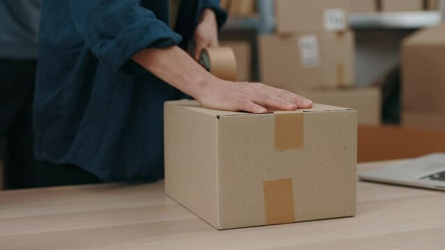 Cropped view of the caucasian man standing at the table and packing parcels with scotch tape attentively. Post service and small business concept.