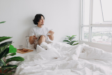 Asian man reading book on the bed with breads and coffee in his apartment.