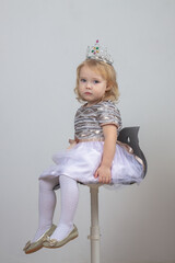 A beautiful baby girl 2 years old in a silver crown and a shiny dress is sitting on a chair on a...