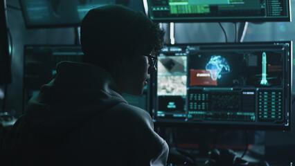 Young male hacker in hat and glasses using computers to hack nuclear warhead and start war while sitting at table in dark room