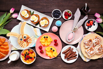 Fun Easter breakfast or brunch table scene. Overhead view on a dark wood background. Bunny pancake, egg nests, chick fruit and a variety of spring food items.