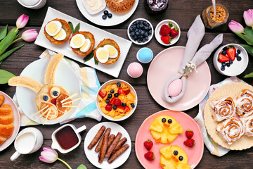 Fun Easter breakfast or brunch table scene. Top down view on a dark wood background. Bunny pancake, egg nests, chick fruit and assorted spring food items. - 486762378
