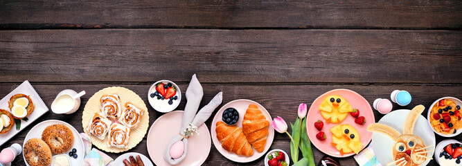 Fun Easter breakfast or brunch bottom border. Top view on a dark wood banner background. Bunny pancake, egg nests, chick fruit and a variety of spring food items. Copy space.