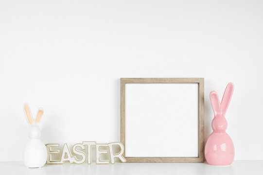 Mock up wood frame with Easter decorations on a white shelf. Modern glass bunny and wooden Easter sign. Square frame against a white wall. Copy space.
