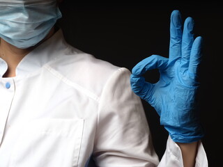 female doctor in a coat and mask shows ok with a gloved hand on a gray background