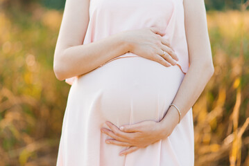 A pregnant woman in a pink delicate dress holds her hands on her stomach, hugs him