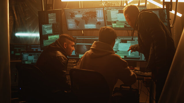 Young man in hoodie helping associate to hack database for criminal while gathering around table in dim room