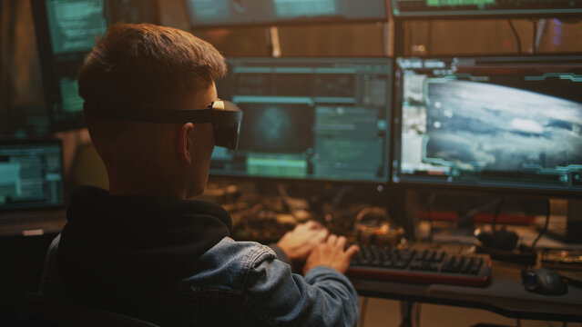 Young man in VR goggles using computer to hack launch of nuclear bomb to start war while sitting in dim room