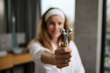 Close-up of a facial massager with two faceted rollers for rejuvenating facial massage in the hands of a blurred red-haired woman in a white bathrobe during her morning routine in the bathroom at home