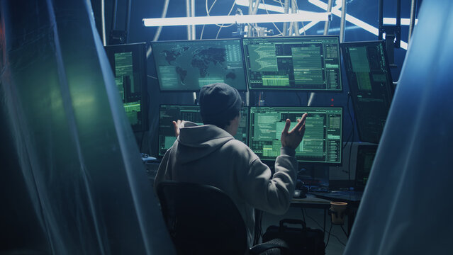 Happy young man in hoodie with glasses and hat dancing in excitement after successful hacker attack, while sitting at desk with computers in dark room