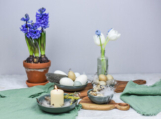 Dekoration Ostern mit Eiern, Federn und Frühlingsblumen auf einem Tisch mit Teller, Schale, Blumenvase und Holzbrett, Frühling dekorieren, Tulpe, Hyazinthe
