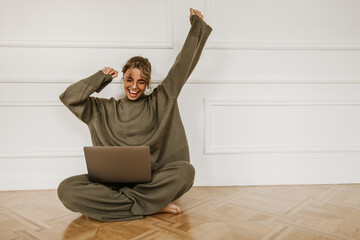 Smiling young caucasian woman using laptop while sitting on floor at home. Beautiful girl with...