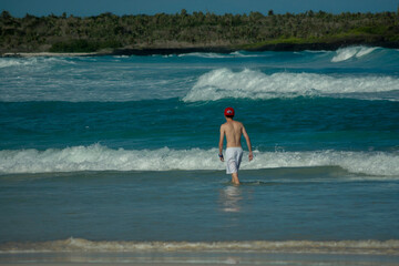 Young man with a red hat facing the turquoise, blue and green sea waves at Tortuga Bay, Galapagos Islands.