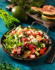 Tropical salad on a bright turquoise background. Fresh salad in a gray plate of arugula, papaya, avocado, cherry tomatoes, strachatella and pine nuts. Exotic dish. Background image, copy space