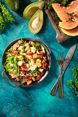 Tropical salad on a bright turquoise background. Fresh salad in a gray plate of arugula, papaya, avocado, cherry tomatoes, strachatella and pine nuts. Exotic dish. Background image, copy space