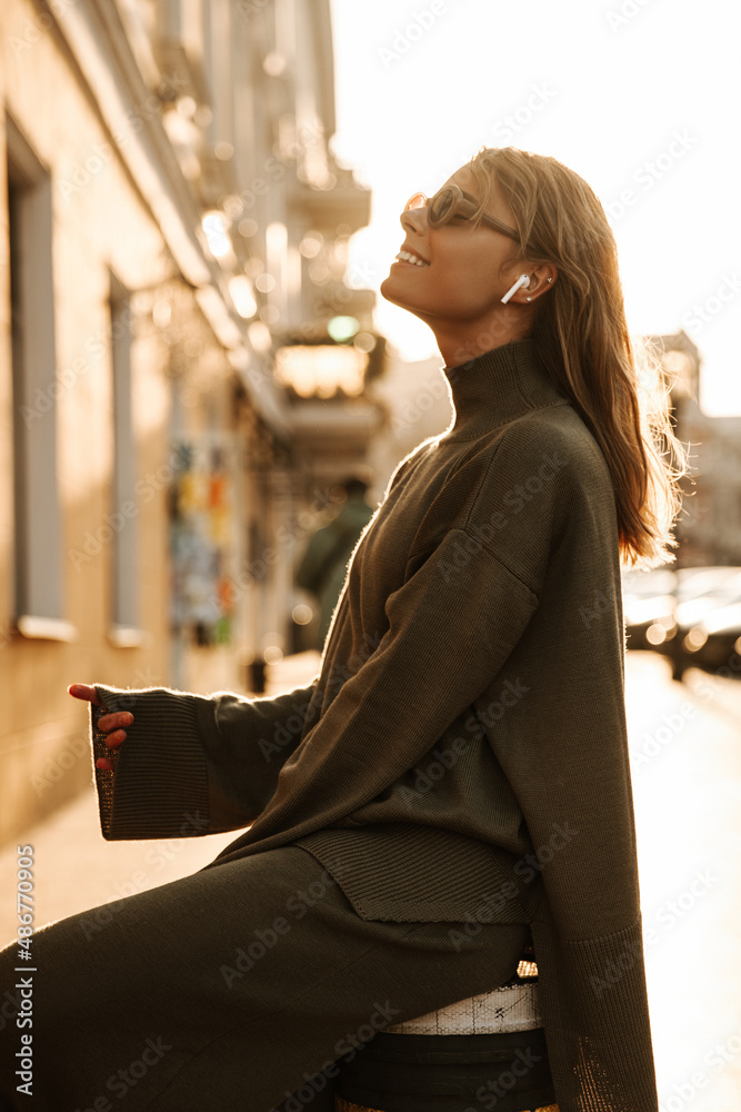 Wall mural Profile photo of cute caucasian young girl enjoying favorite song through headphones on street. Beauty model in grey spacious suit smiles closing her eyes. Autumn trendy fashion, concept.