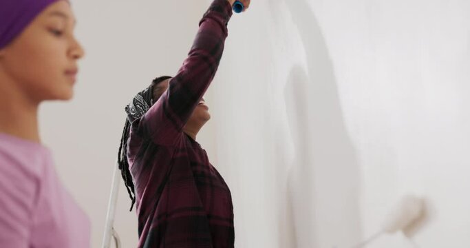 Sweet girl is painting wall with a roller that is covered with white paint together with mom, the woman is cheering her daughter to continue, together they are renovating a room in the apartment