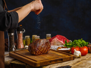 The chef's hands are salting a baked ham on a wooden cutting board. festive dish, restaurant and home cooking recipes, organic food. Dark background.