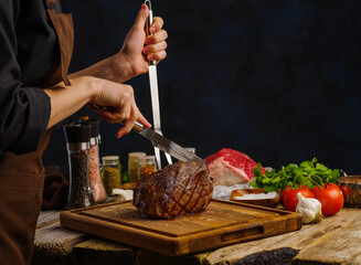The chef slices a juicy ham on a cutting board. Vegetables, herbs, spices. Dark background. Restaurant, hotel, cafe, home cooking, recipes for meat dishes.
