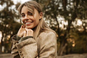 Close-up of radiant young caucasian girl looking at camera on blurred street background. Smiling blonde woman touches her chin, wearing jacket. Outdoor autumn lifestyle happiness emotions