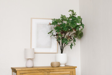 An artificial flower in a pot on a wooden chest of drawers in the living room, decorative items and a frame on the wall in a Scandinavian minimalist style