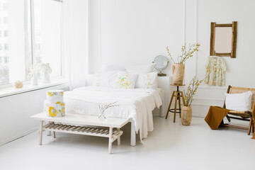 Light airy white with yellow shades bedroom in the house. Cozy bed and vases with spring branches