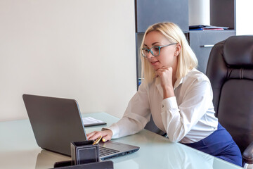 Beautiful caucasian blond haired woman wearing white shirt and blue skirt sits on chair in office and works using laptop. Conference call theme.