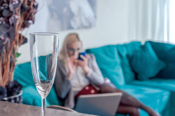 Blurred caucasian blond haired woman in eyeglasses sits on blue sofa and holds smartphone. Woman wears white shirt, grey jacket, skirt and stockings. Focus on foreground with empty wine glass.