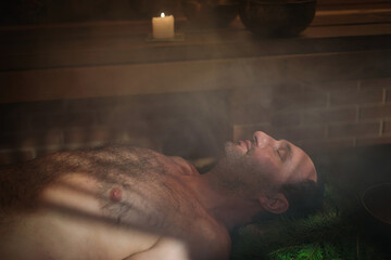 A man lies on spruce branches in a sauna, he is being performed a Tibetan ritual.