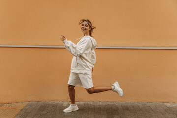 Active young caucasian lady with flying hair runs along street wall laughing at camera. Pretty sportswoman in white hoodie, shorts and shoes. Energetic lifestyle concept