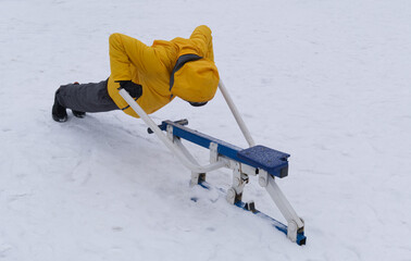 An elderly man in a yellow jacket is engaged in recreational gymnastics on exercise equipment outdoors in a natural park in winter against the backdrop of snow.