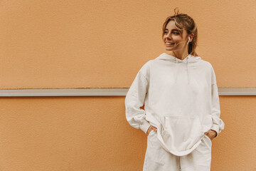 Medium photo cute young caucasian woman with smile on face looking into distance against backdrop with space for text. Blonde with gathered hair holds hands in pockets. Lifestyle, beauty concept