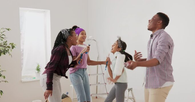 Crazy happy family dances in middle of room during break from painting the walls, a woman and her daughters sing to the rollers, a man pretends to play the guitar, fun apartment renovation