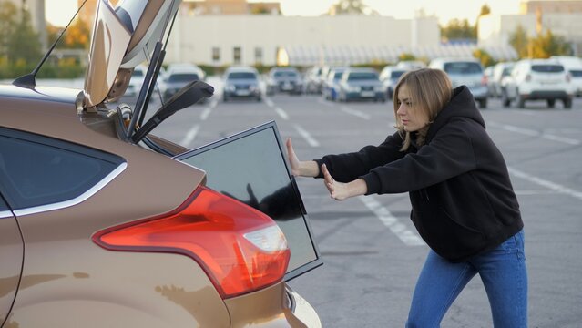 Woman funny trying to shove purchased modern tv in car trunk at supermarket parking, delivery concept
