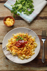 Pasta bolognese garnished with greens and cheese in a plate on a wooden table on a glossy board next to a fork and greens.