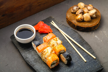 Assorted rolls on a black board next to sticks, soy sauce and baked mushrooms on a dark background.