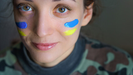 Indoor portrait of young girl with blue and yellow ukrainian flag on her cheek wearing military...