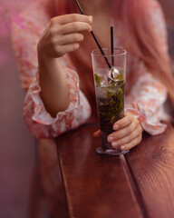 A girl is drinking a refreshing drink with mint through a straw from a glass. A customer at a table in a summer cafe. Woman's hands close up. Pink colors.