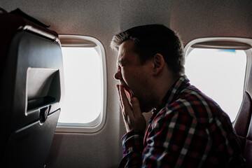 a man in a plaid shirt sits by the window of the plane and yawns. long flight. 