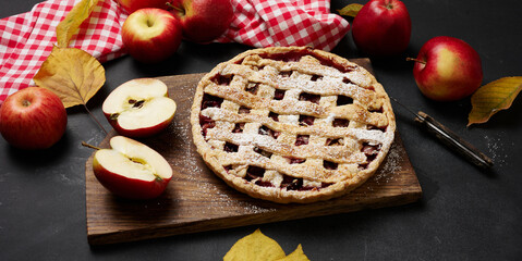 baked round traditional apple pie on brown wooden board and fresh red apples, top view