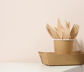 paper cardboard brown plates and cups, wooden forks and knives on a white table, beige background....
