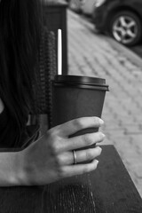 Girl with a ring on her finger holds a cup with a white tube at a table in a city cafe. Black and white photo
